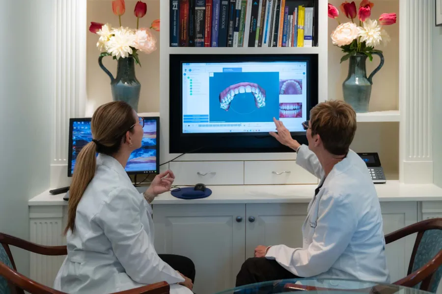 Two dentists in white coats discuss dental scan images on a computer screen in a bright office with flowers and books.