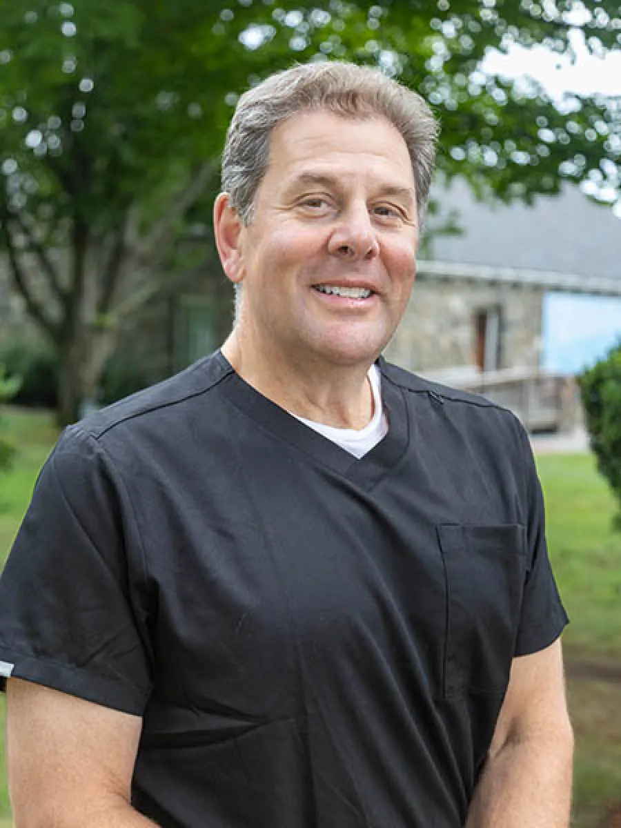 Smiling middle-aged man in black scrub top stands outdoors with trees and stone building in background.