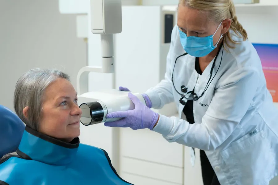 Dentist wearing mask and gloves prepares X-ray machine for elderly female patient in dental clinic.