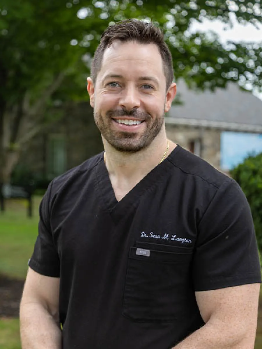 Smiling male healthcare professional in black scrubs standing outdoors with green trees and buildings in the background