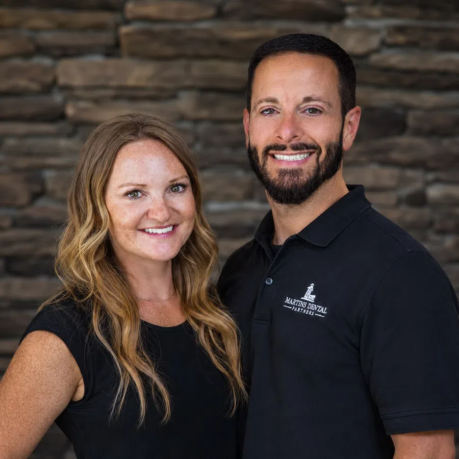 Smiling man and woman in black shirts pose together against a stone wall background, showing friendly expressions.