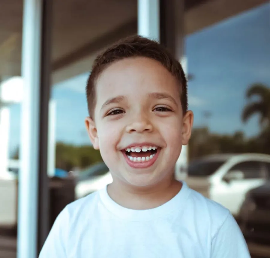 Smiling young boy with short hair wearing a white shirt standing outdoors with blurred cars and palm trees in background
