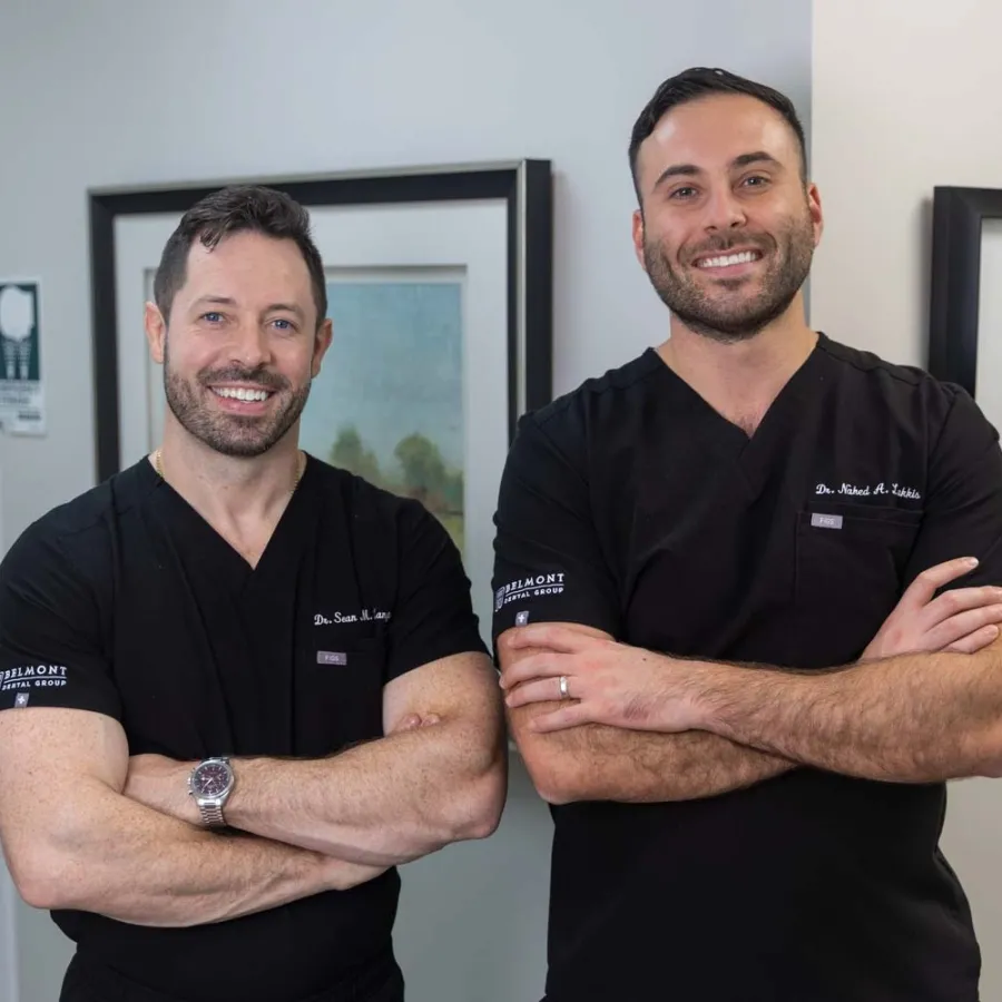 Two male dentists in black scrubs smiling with arms crossed inside a dental office with framed art.