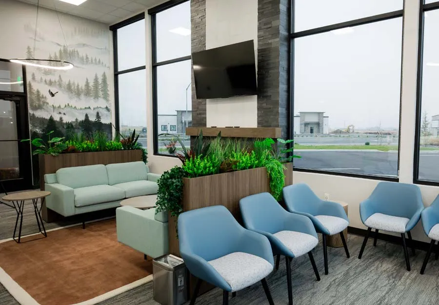 Modern waiting room with blue chairs, green plants, large windows, TV screen, and nature-themed wall mural.