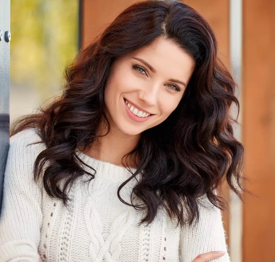 Smiling woman with long wavy brown hair wearing a white knitted sweater standing by a window.