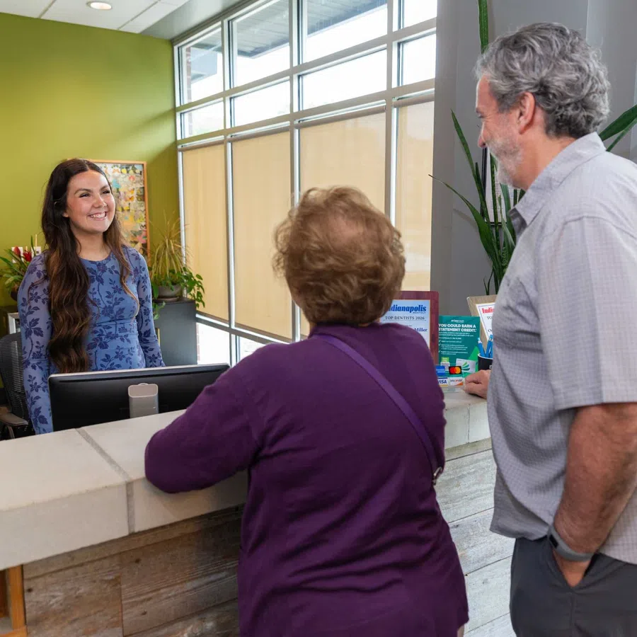 A receptionist smiling and assisting two elderly visitors at a modern office reception desk.