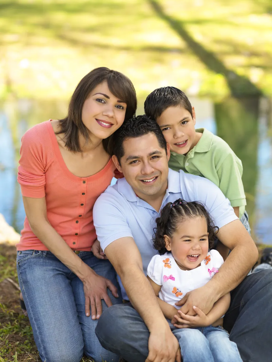 Smiling family of four sitting outdoors by a pond on a sunny day enjoying quality time together.