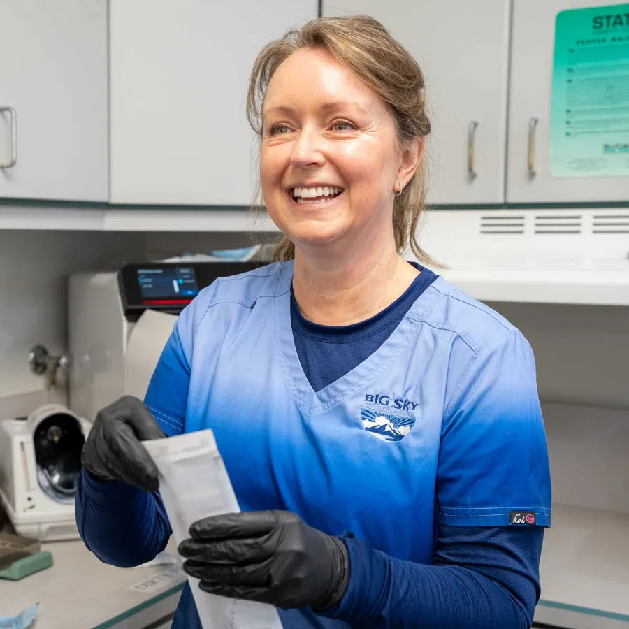 Smiling medical professional in blue scrubs and black gloves holding a sample bag in a clinical lab setting.