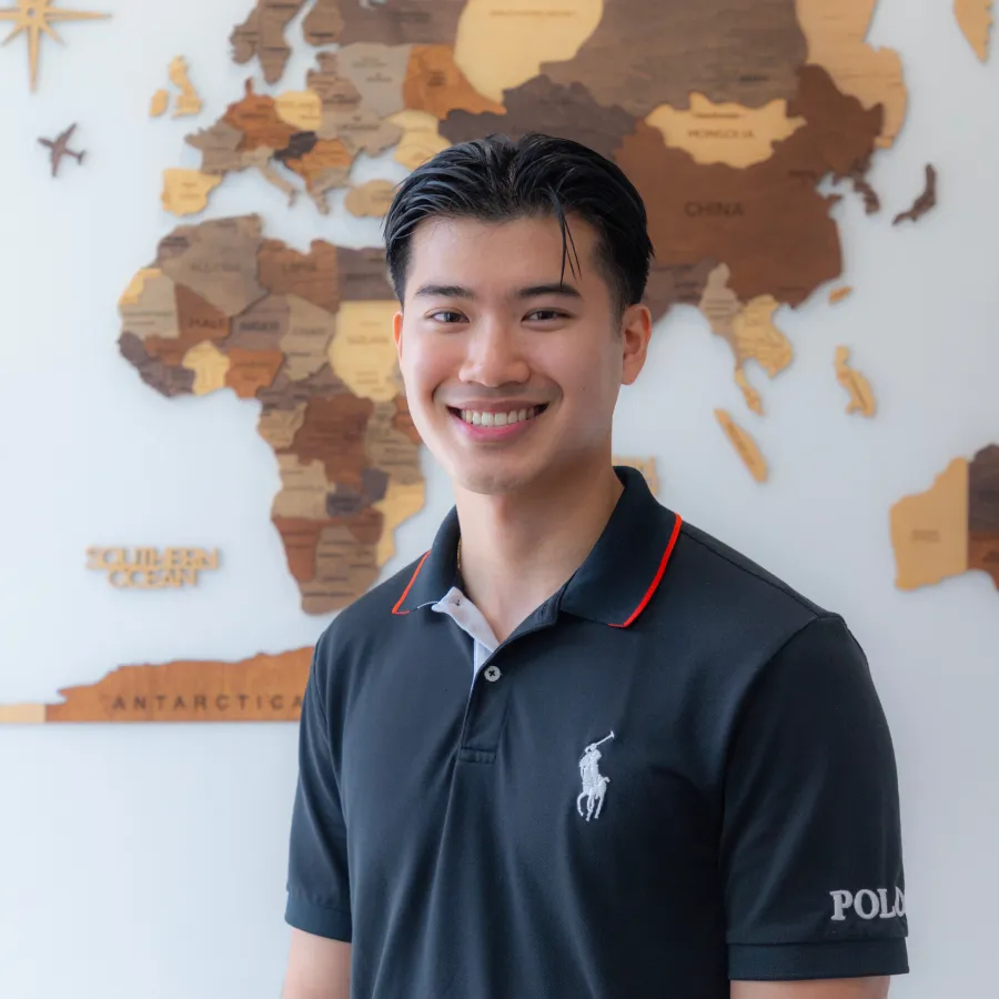 Smiling young man in black Polo shirt standing in front of a wooden world map on white wall