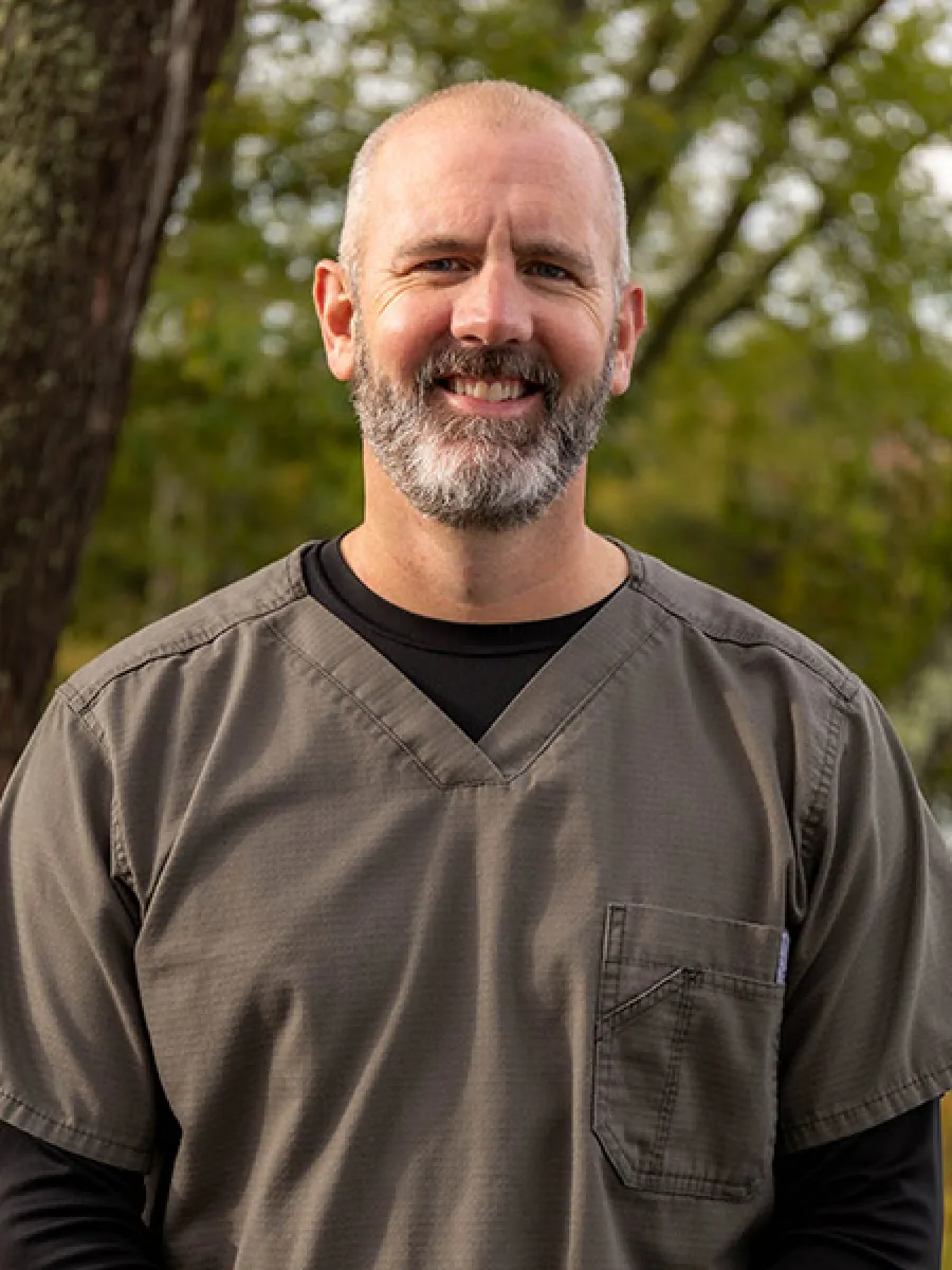 Smiling middle-aged man with a beard wearing gray medical scrubs outdoors with trees in the background