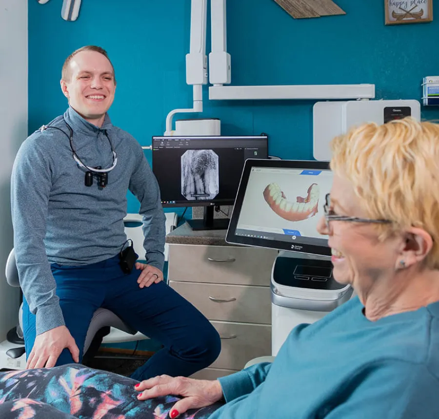 Dentist consulting a smiling senior woman with dental x-rays and 3D teeth model on screens in clinic.