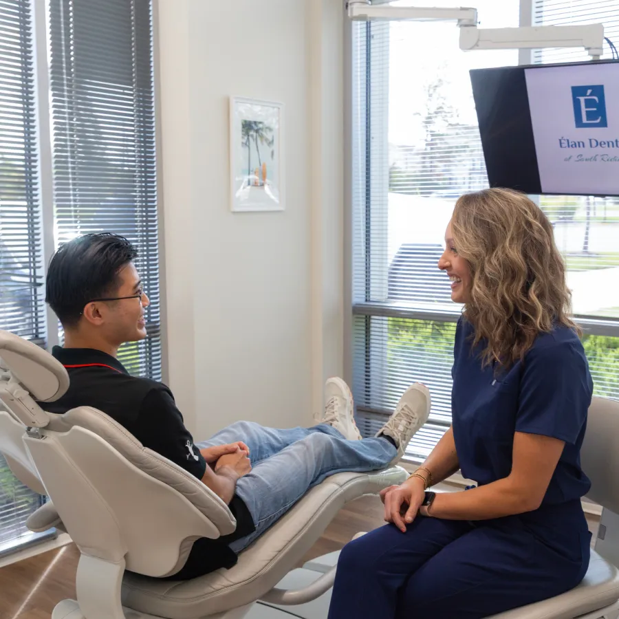 Dentist in navy scrubs smiling and talking with relaxed male patient in dental chair in bright office
