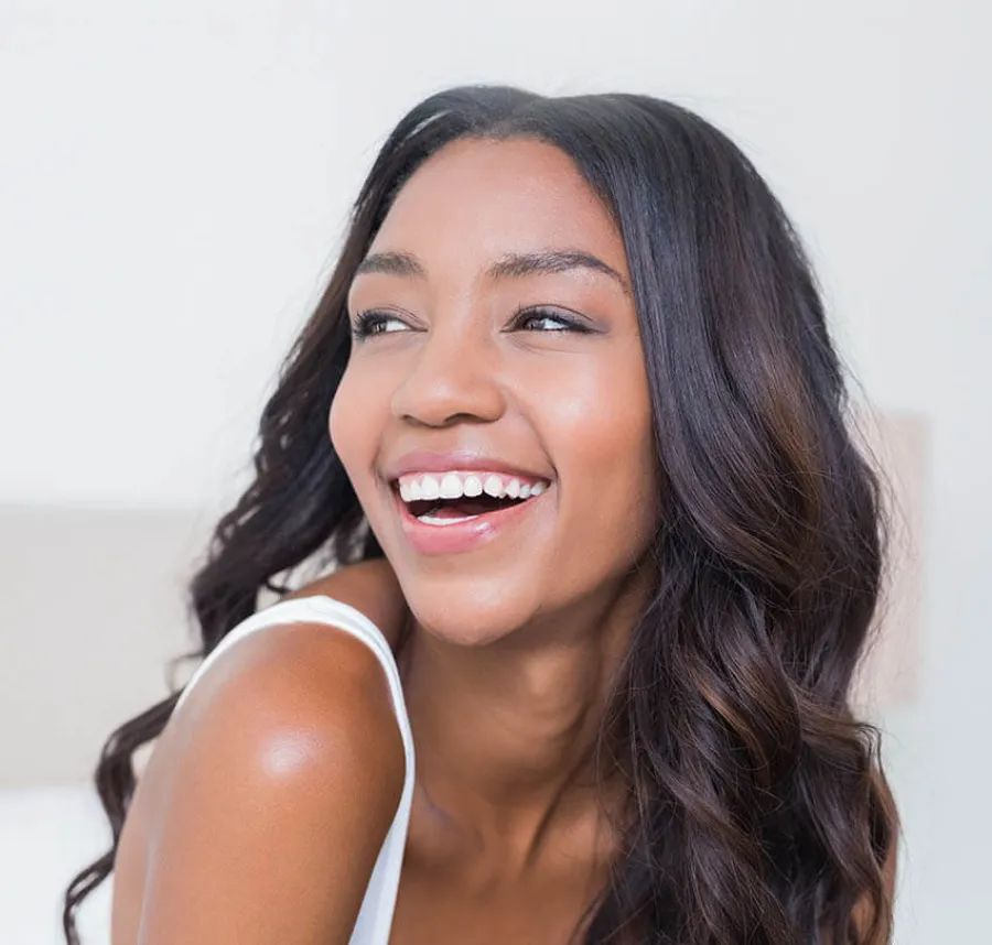 Close-up of a smiling young woman with long wavy hair wearing a white sleeveless top against a light background