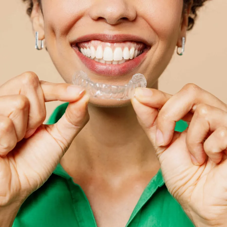 Smiling person holding a clear dental aligner in front of their bright white teeth and green shirt.
