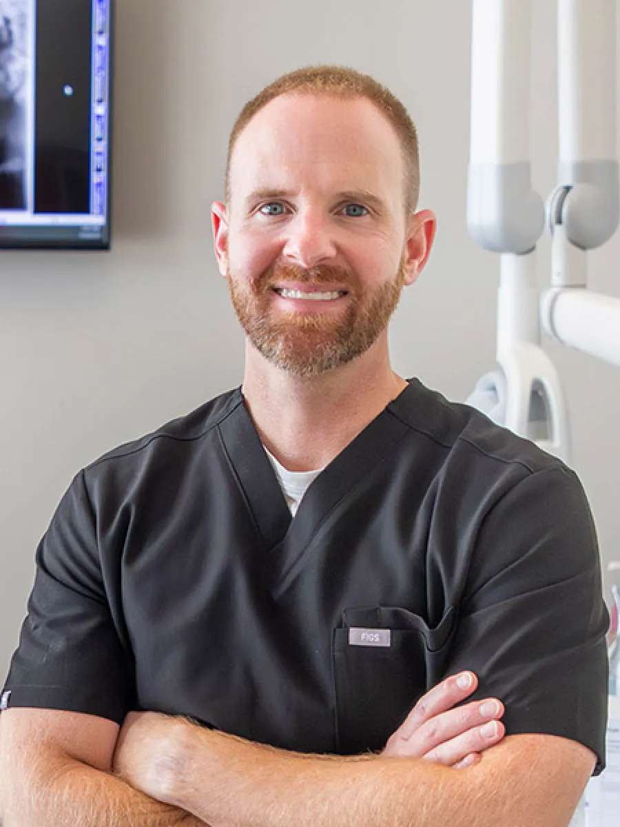 Smiling male dentist with beard in black scrubs standing confidently in dental office with x-ray monitor.