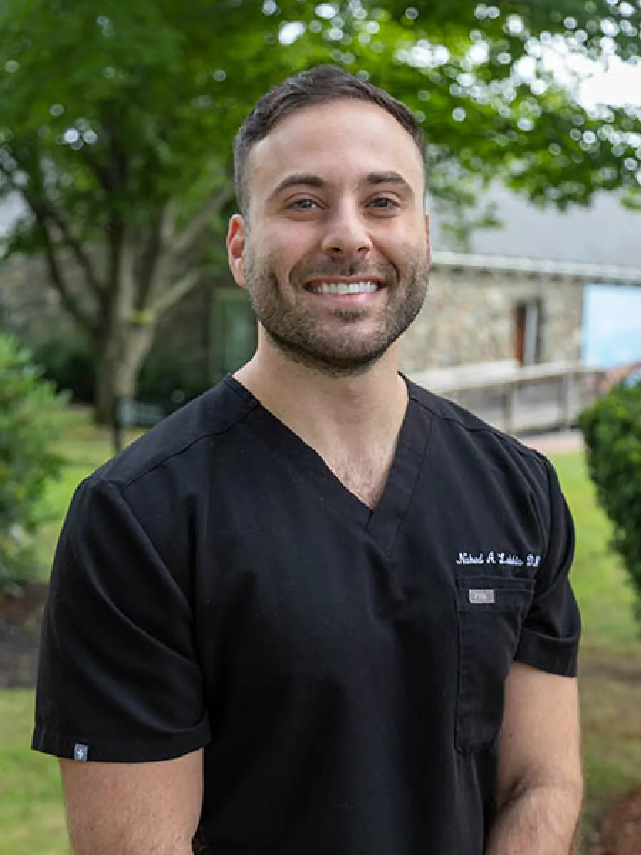 Smiling man in black medical scrubs standing outdoors with greenery in the background