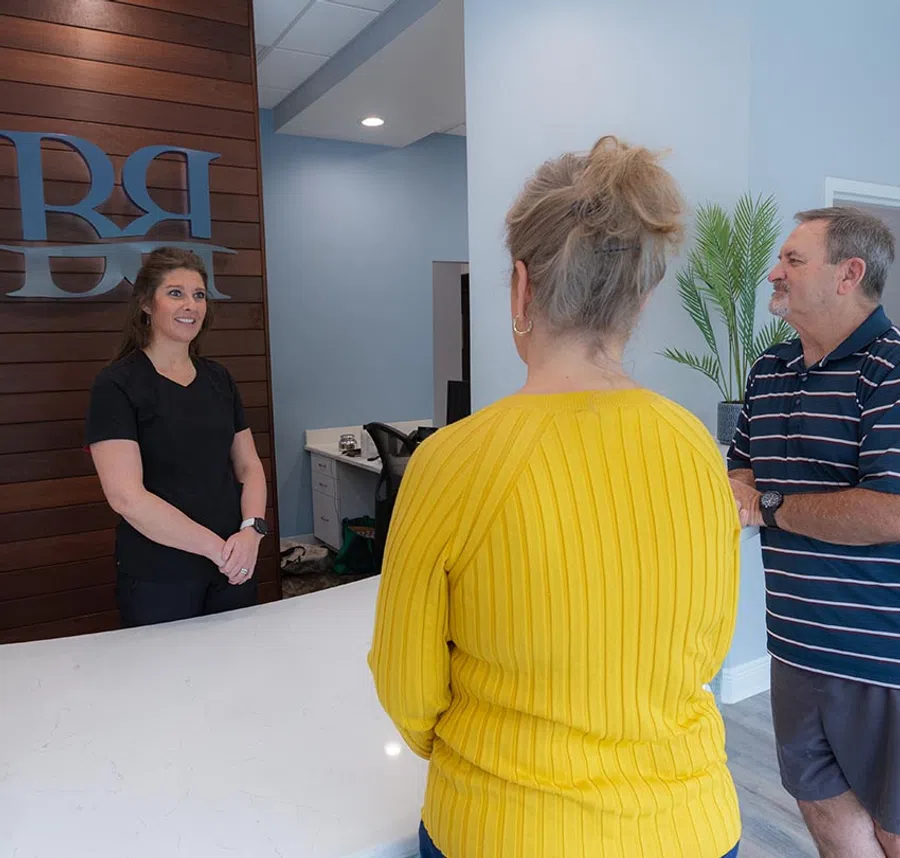 Receptionist in black talking to two visitors at a modern office front desk with wooden accent wall.