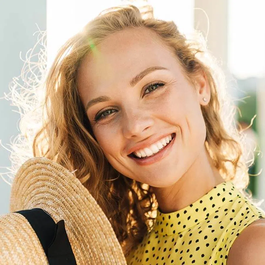 Smiling young woman with dark hair in a white shirt standing outdoors in soft focus background.