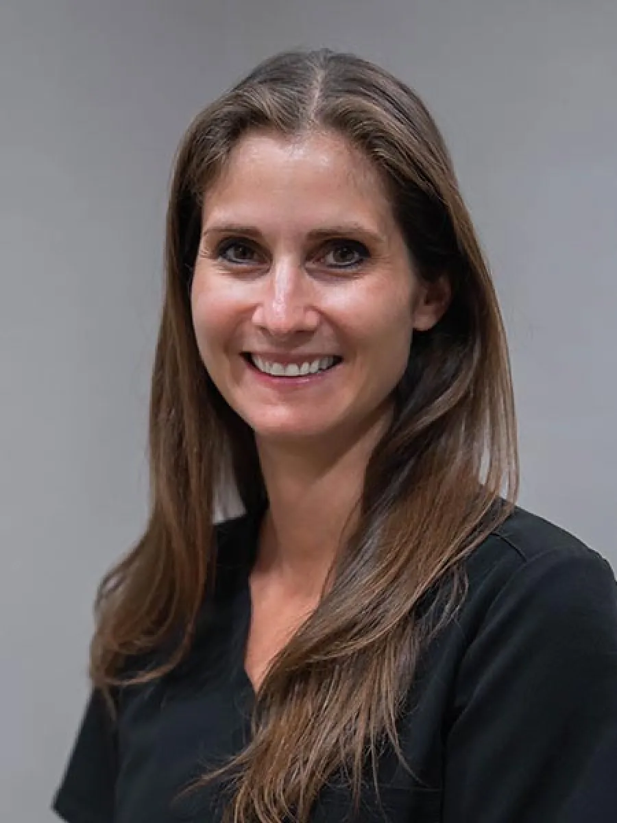 Smiling woman with long brown hair wearing a black top against a plain gray background.