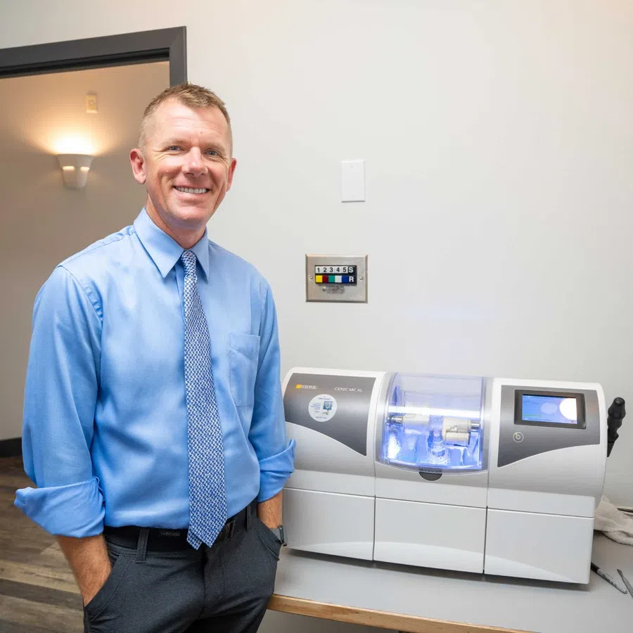 Smiling man in blue shirt and tie standing next to dental milling machine in clinic room.