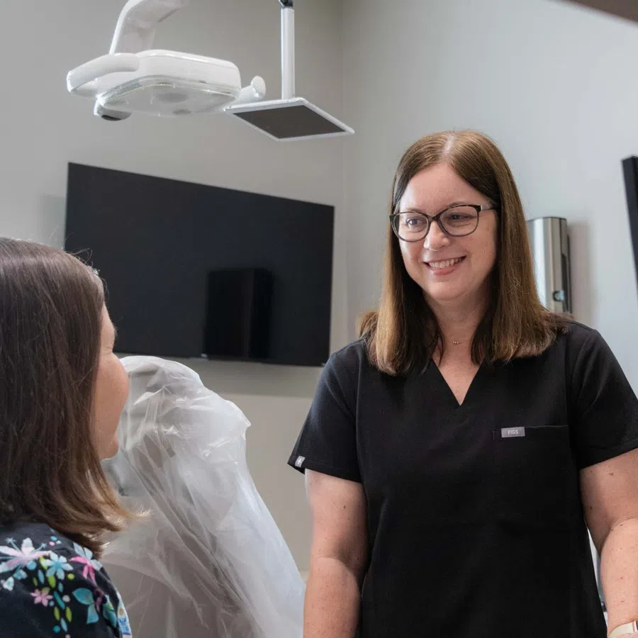 Dentist in black scrubs smiling and talking to a patient in a modern dental office with screens and equipment.