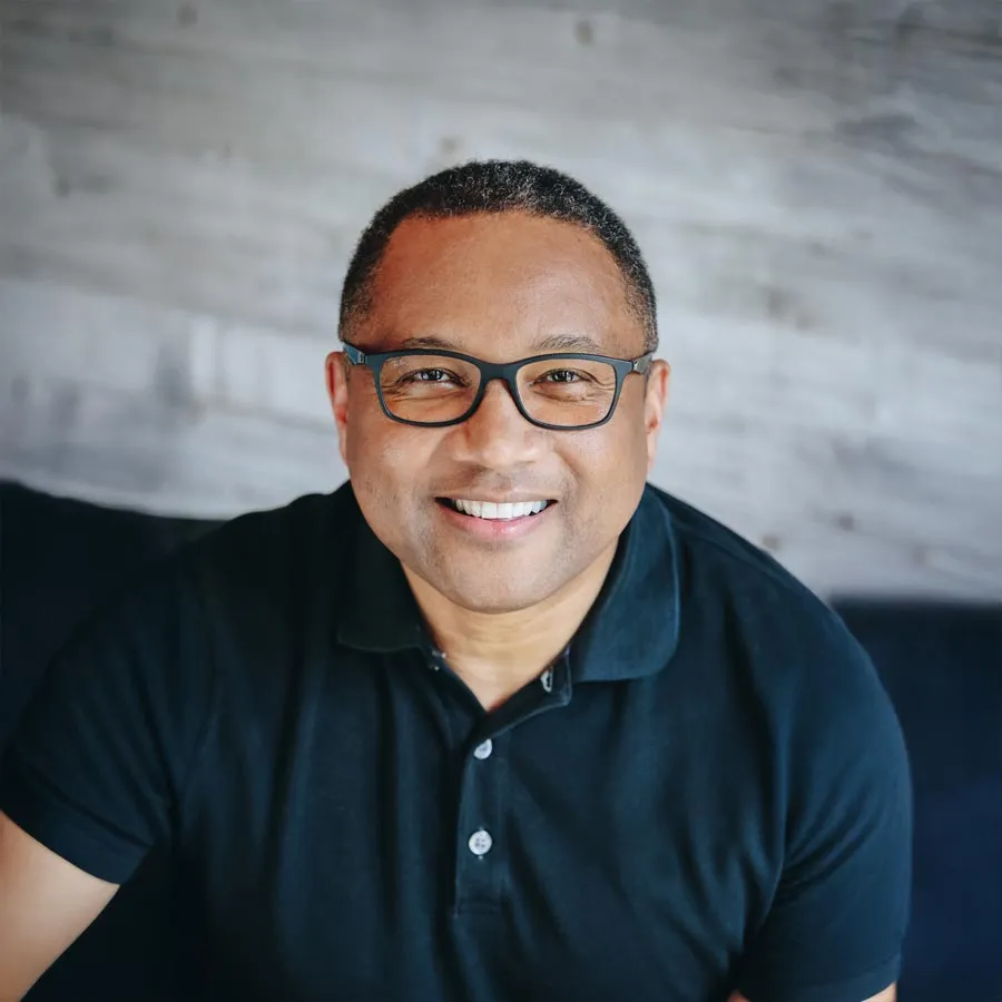 Smiling man in a black polo shirt posing against a rustic wood background.