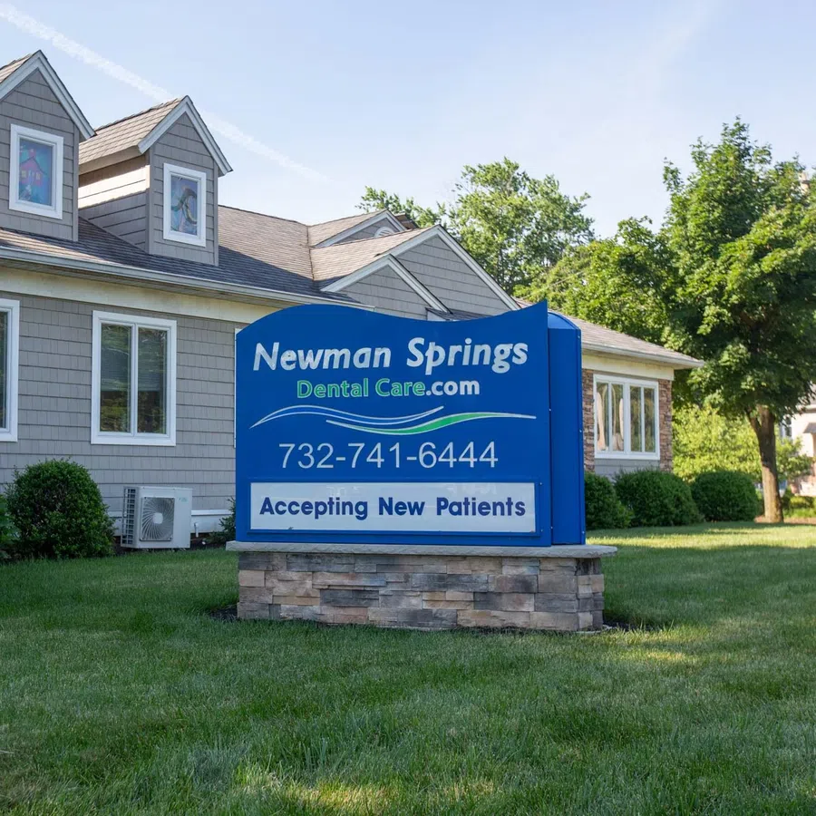 Newman Springs Dental Care office building with a blue sign accepting new patients outside on a grassy lawn.