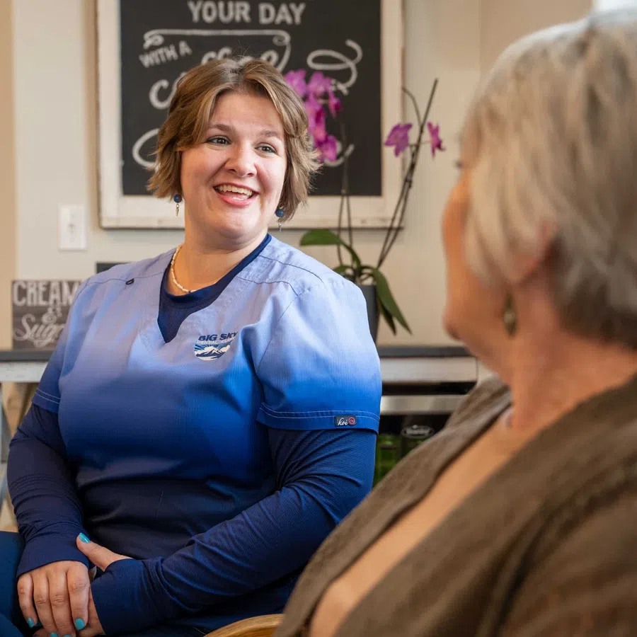 Healthcare worker in blue scrubs smiling and talking with an older woman in a cozy room with a plant and sign