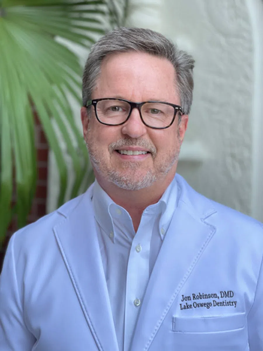 Middle-aged man with glasses wearing a white dental coat standing in front of greenery and white wall.