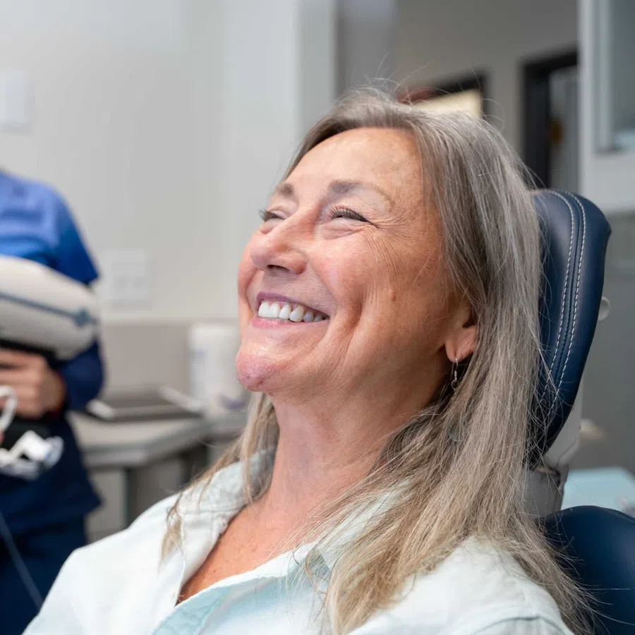 Smiling senior woman sitting in dental chair with dental assistant holding cure light in background