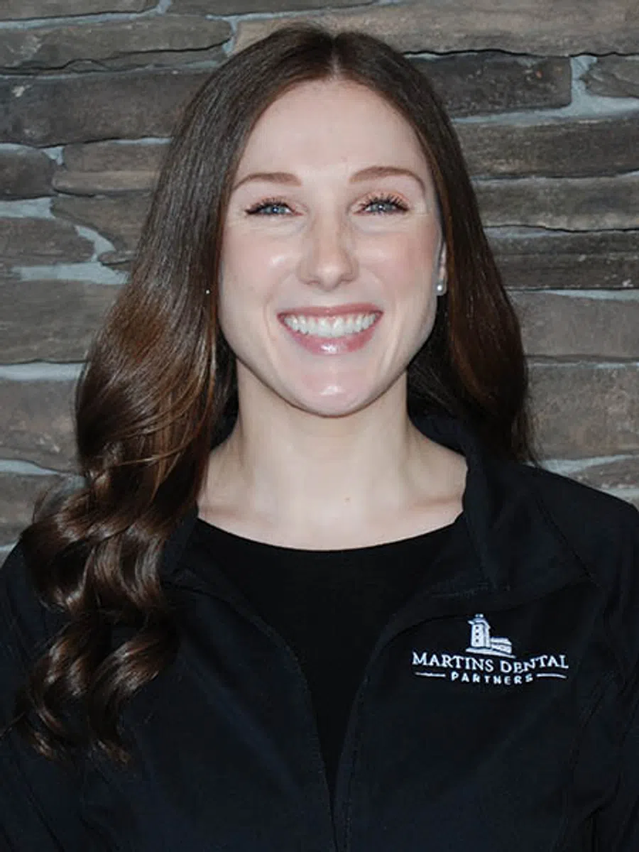 Smiling woman with long brown hair wearing a black Martins Dental Partners jacket against a stone wall.