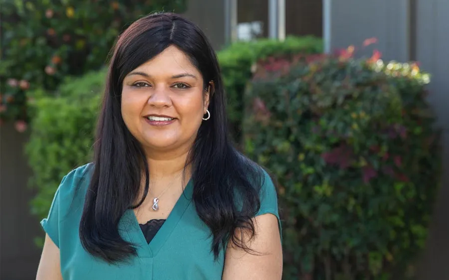 Smiling woman with long black hair wearing green top and silver necklace standing outdoors with green foliage background