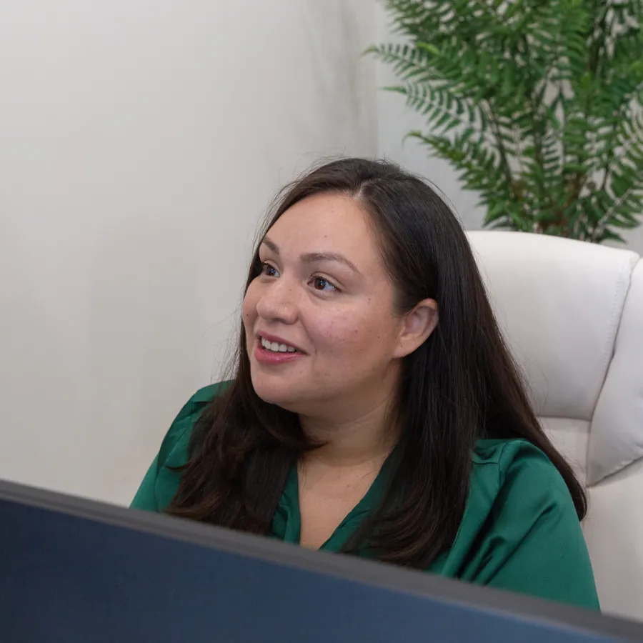 Woman with long dark hair in a green blouse smiles while sitting at a desk with a computer screen in front of her.