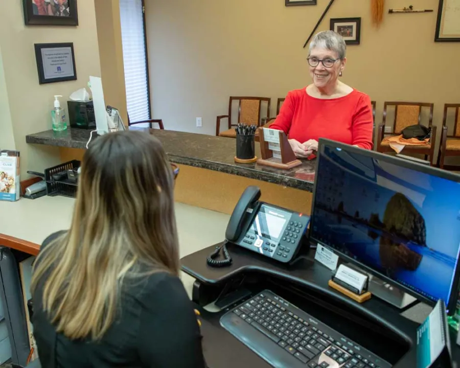 Older woman in red shirt smiling at receptionist across the counter in a professional office setting.