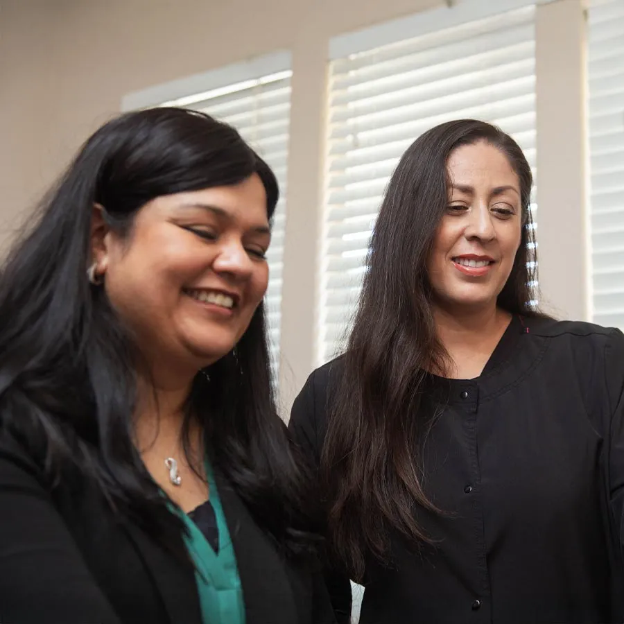 Two women smiling and interacting indoors with white blinds in the background, dressed in black and green clothing.