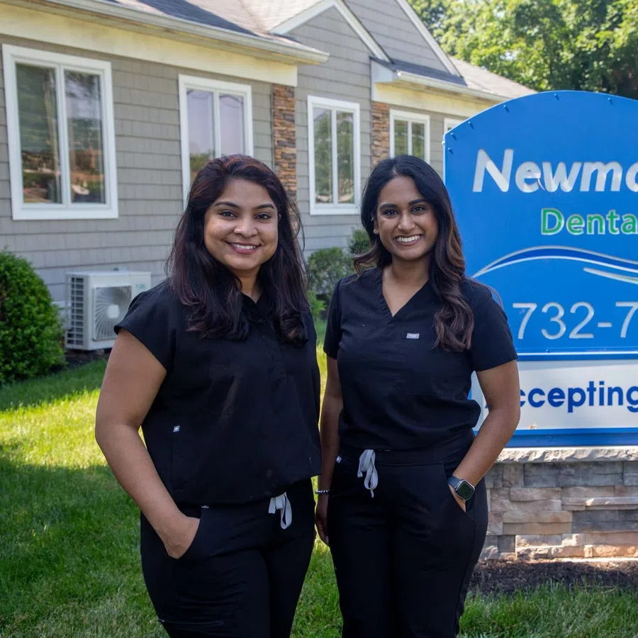 Two smiling women in black scrubs standing outside near a dental office sign on a sunny day.