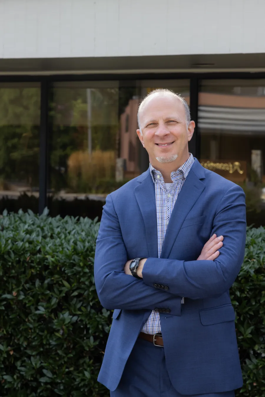 Smiling man in blue suit standing outdoors with arms crossed in front of green bushes and a building window.