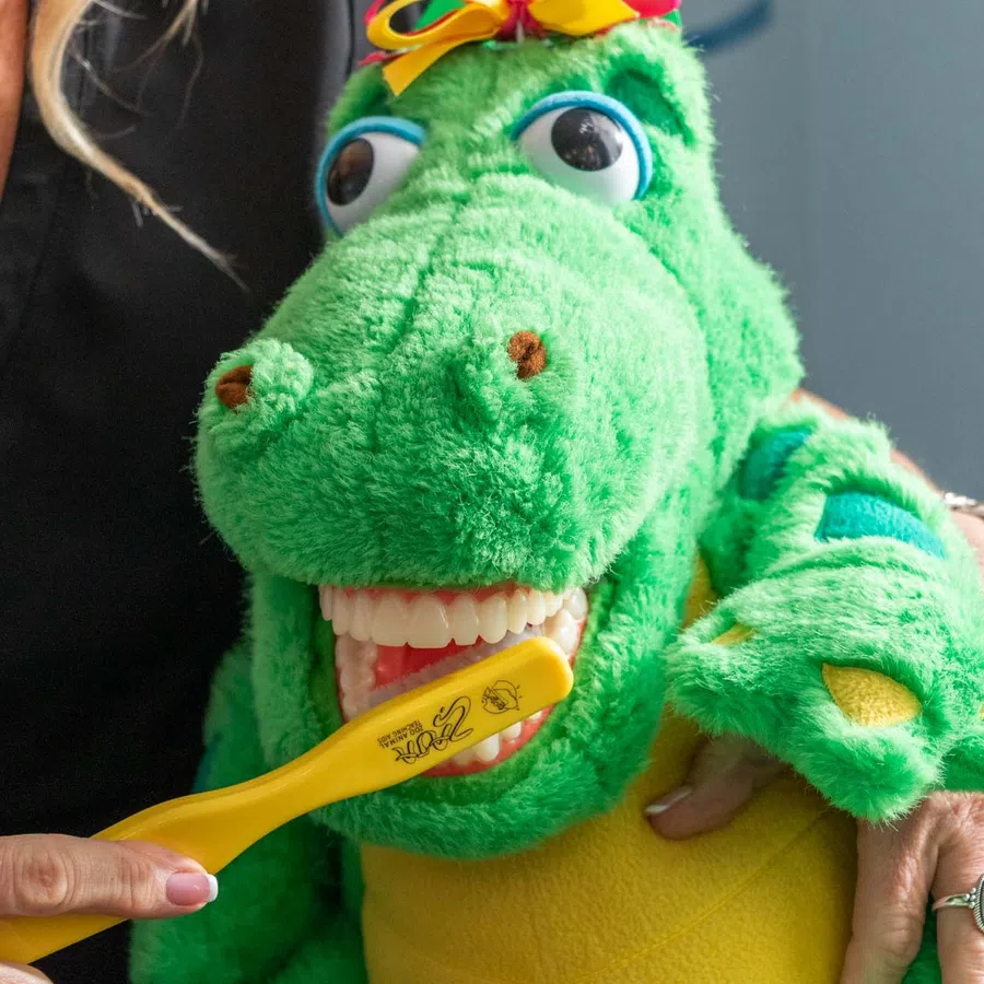 Person uses a yellow toothbrush to brush the teeth of a green dinosaur puppet wearing a bow on its head.