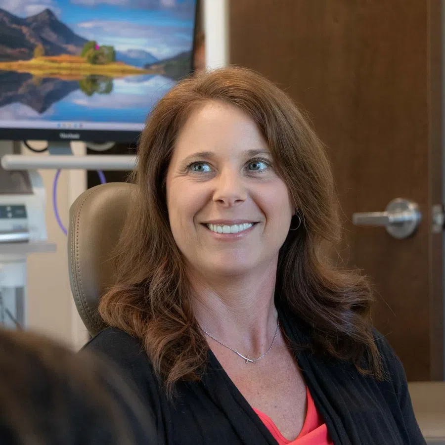 Smiling woman with brown hair sitting in a dental chair during a consultation with a blurred person.