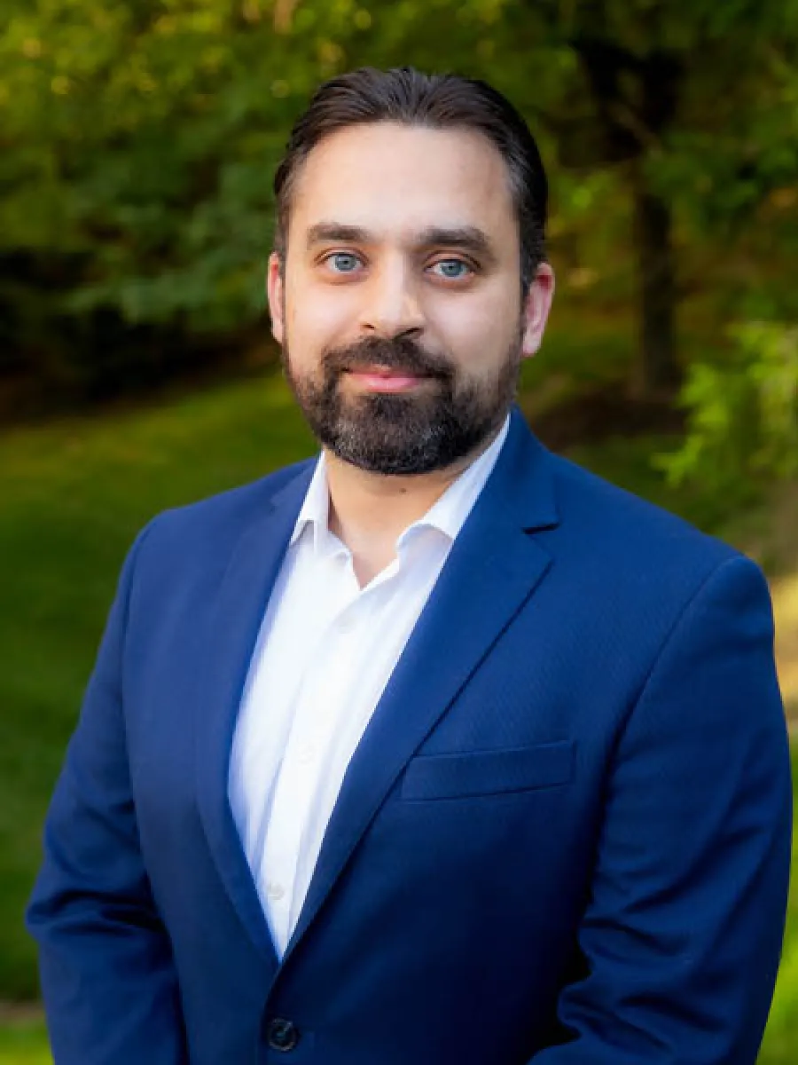 Smiling man with beard wearing a blue suit and white shirt outdoors with green trees in the background