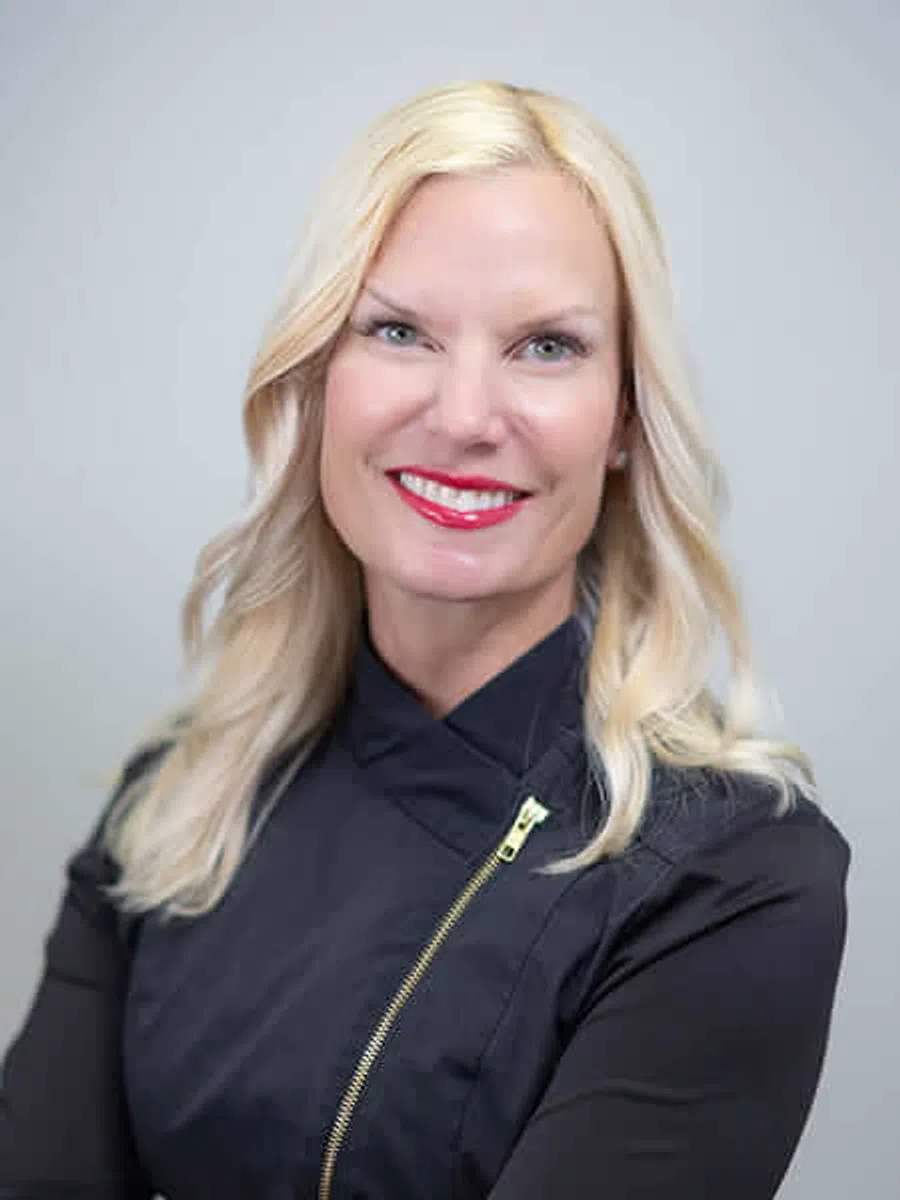 Smiling blonde woman with wavy hair, red lipstick, and black zip-up top against a light grey background