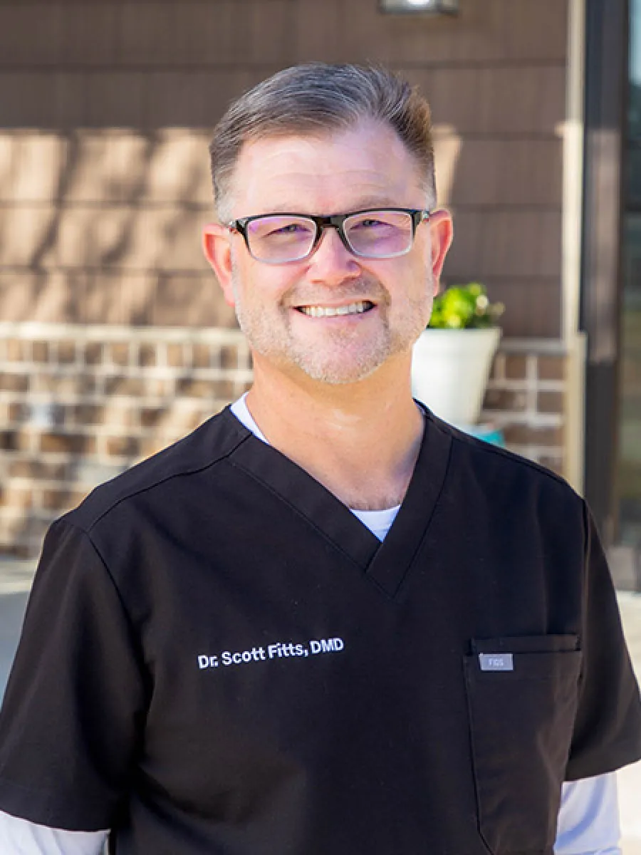 Smiling male dentist wearing black scrubs with name tag Dr. Scott Fitts standing outdoors with brick wall background.