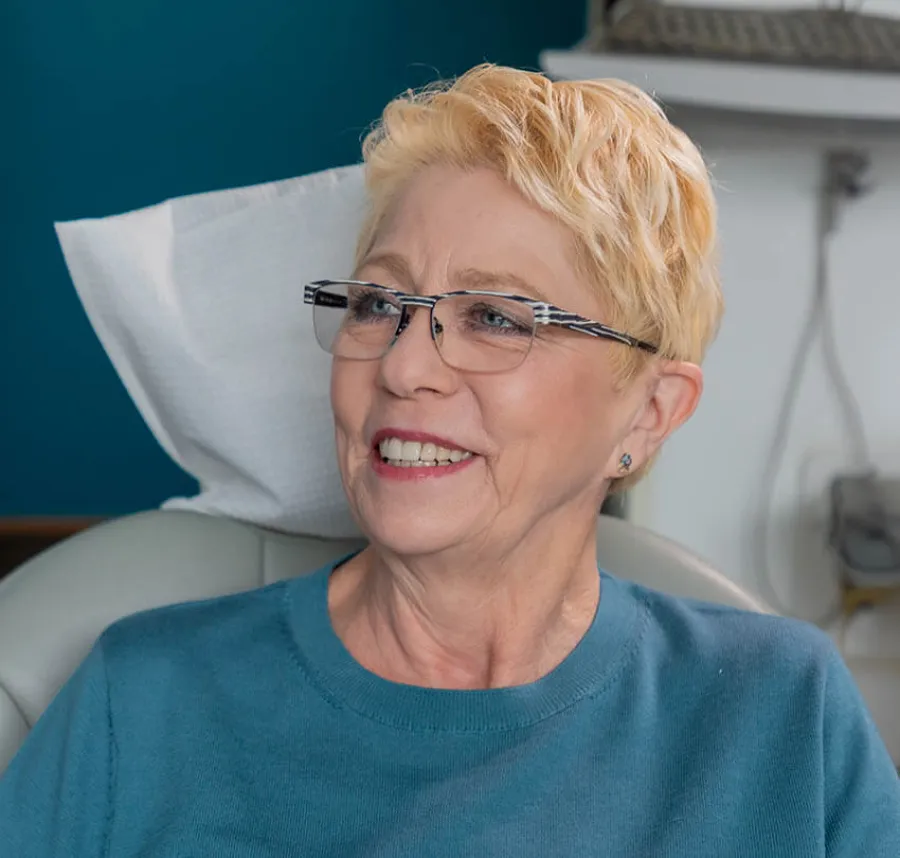 Smiling elderly woman with short blonde hair and glasses sitting in a medical or dental chair, wearing a teal sweater.