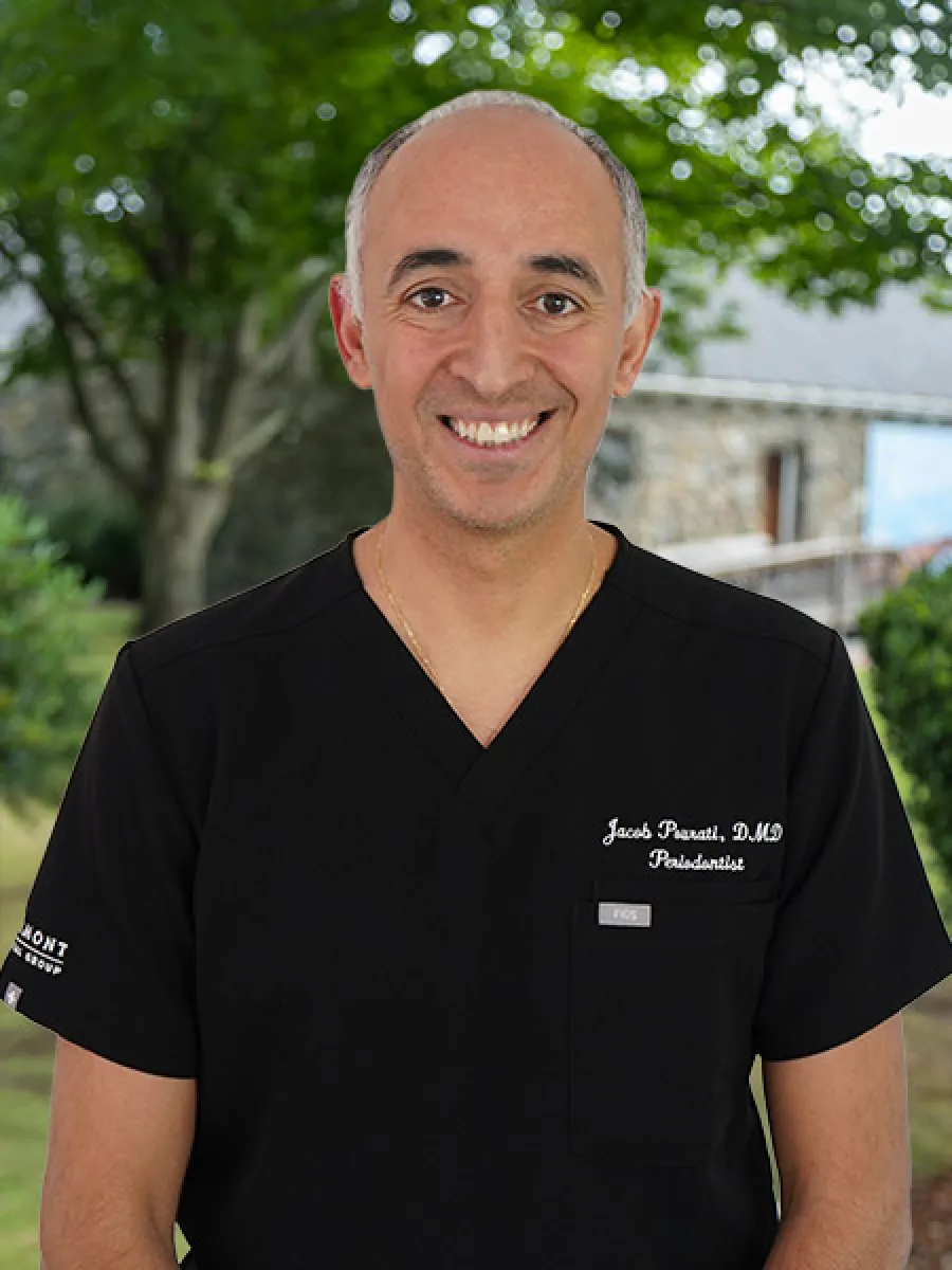 Smiling male doctor wearing black scrubs outside with greenery and stone building background.