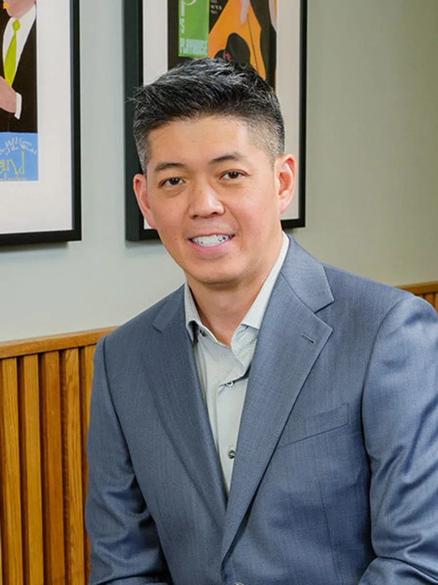 Smiling man in blue suit sitting indoors with framed artwork and wooden paneling in the background.