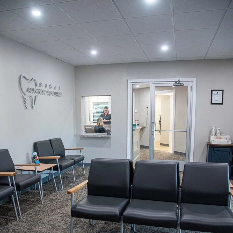 Modern dental office waiting room with black chairs, gray carpet, and reception windows with staff inside.