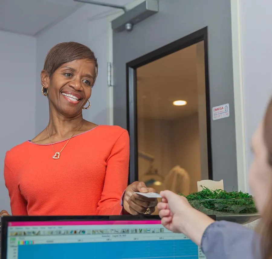 Smiling woman in orange shirt hands a card to person at computer in office reception area.