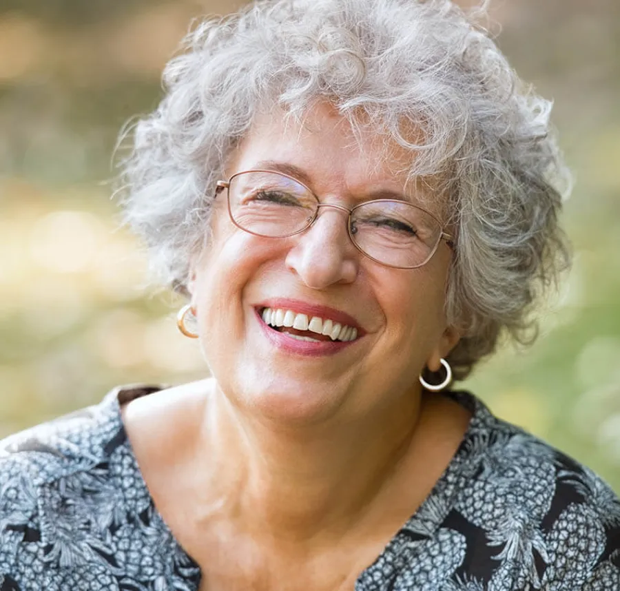 Smiling elderly woman with curly gray hair, glasses, and hoop earrings in an outdoor setting.