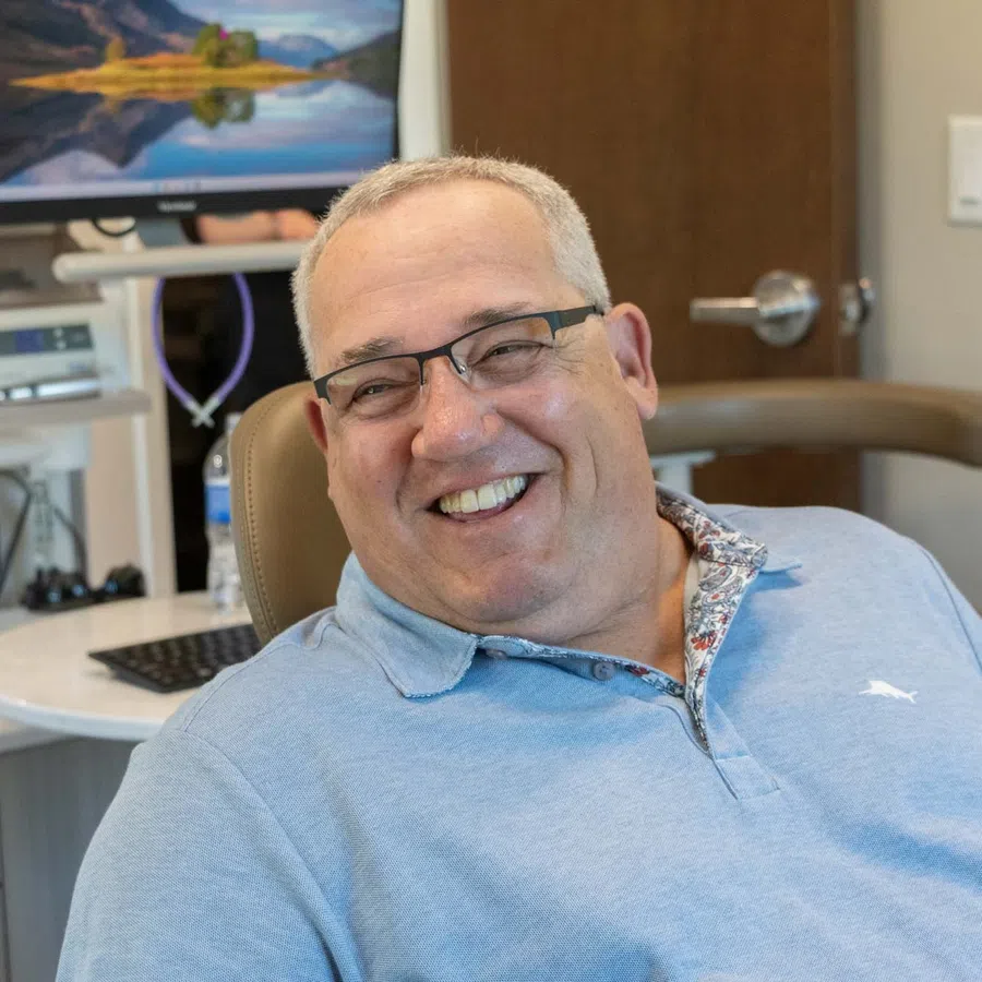 Smiling middle-aged man wearing glasses sitting in a dental chair in a modern clinic with computer screen in background