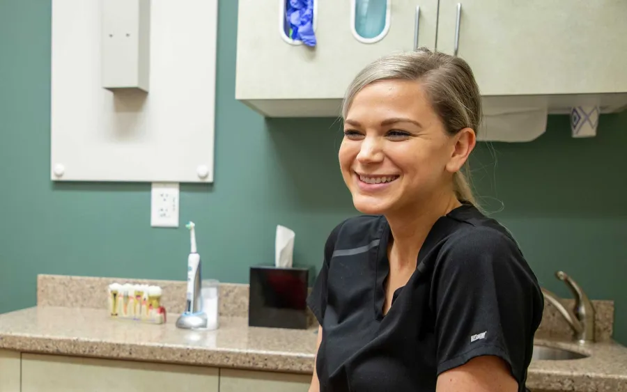 Smiling female dental professional in black scrubs standing in dental office with tools and supplies in background